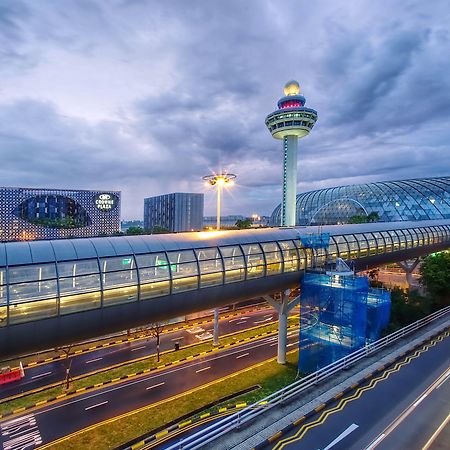 Crowne Plaza Changi Airport By Ihg Σιγκαπούρη Εξωτερικό φωτογραφία The photo depicts an airport scene, likely featuring a modern terminal structure with a distinctive, futuristic design. In the foreground, there is a pedestrian bridge or walkway, along with roads leading to the terminal. A control tower can be seen 