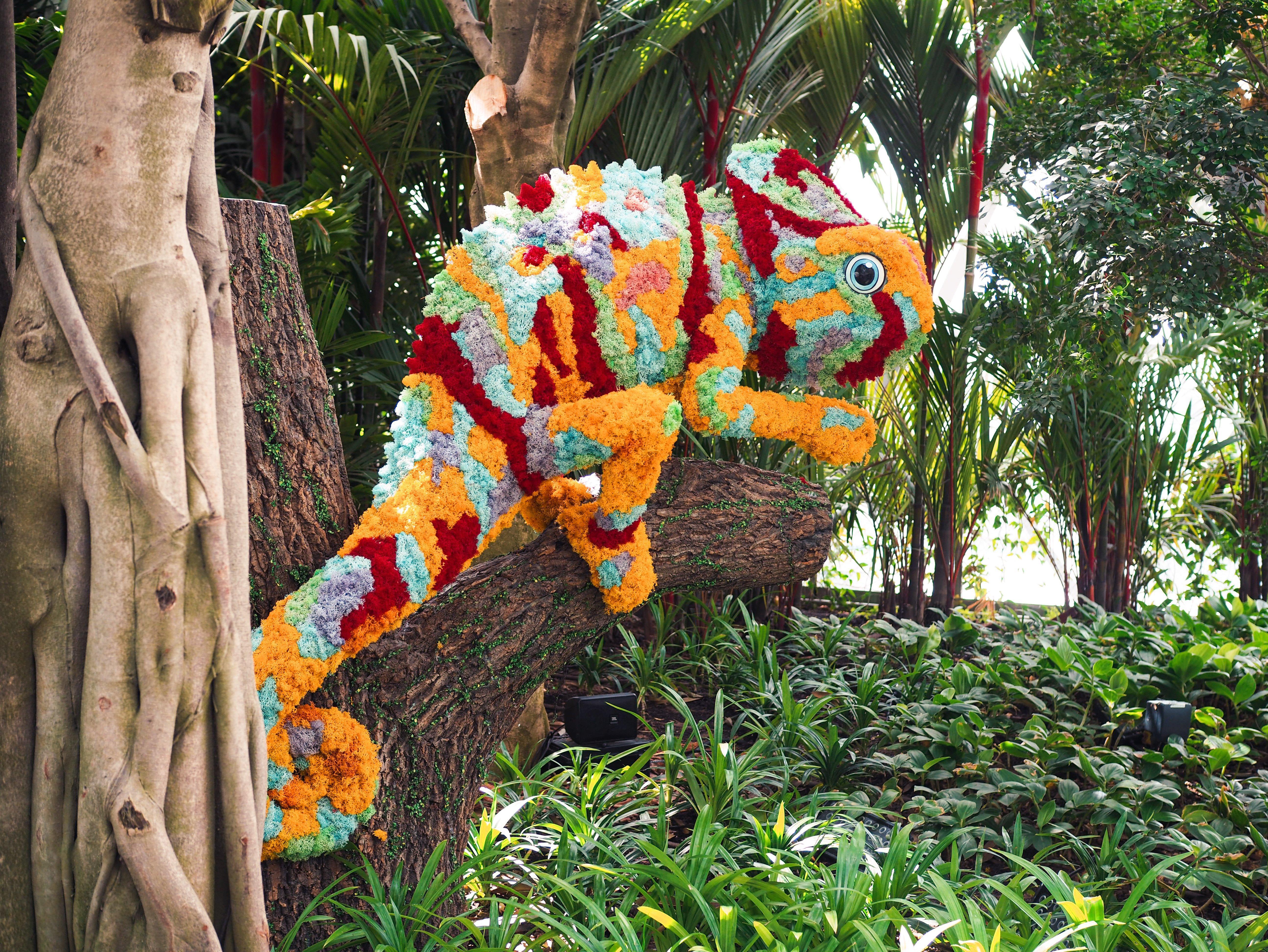 Crowne Plaza Changi Airport By Ihg Σιγκαπούρη Εξωτερικό φωτογραφία The photo shows a vibrant and colorful chameleon sculpture made from flowers and plant materials. The chameleon is positioned on a tree trunk, blending in with the lush greenery of its surroundings. The colors of the sculpture include shades of orang