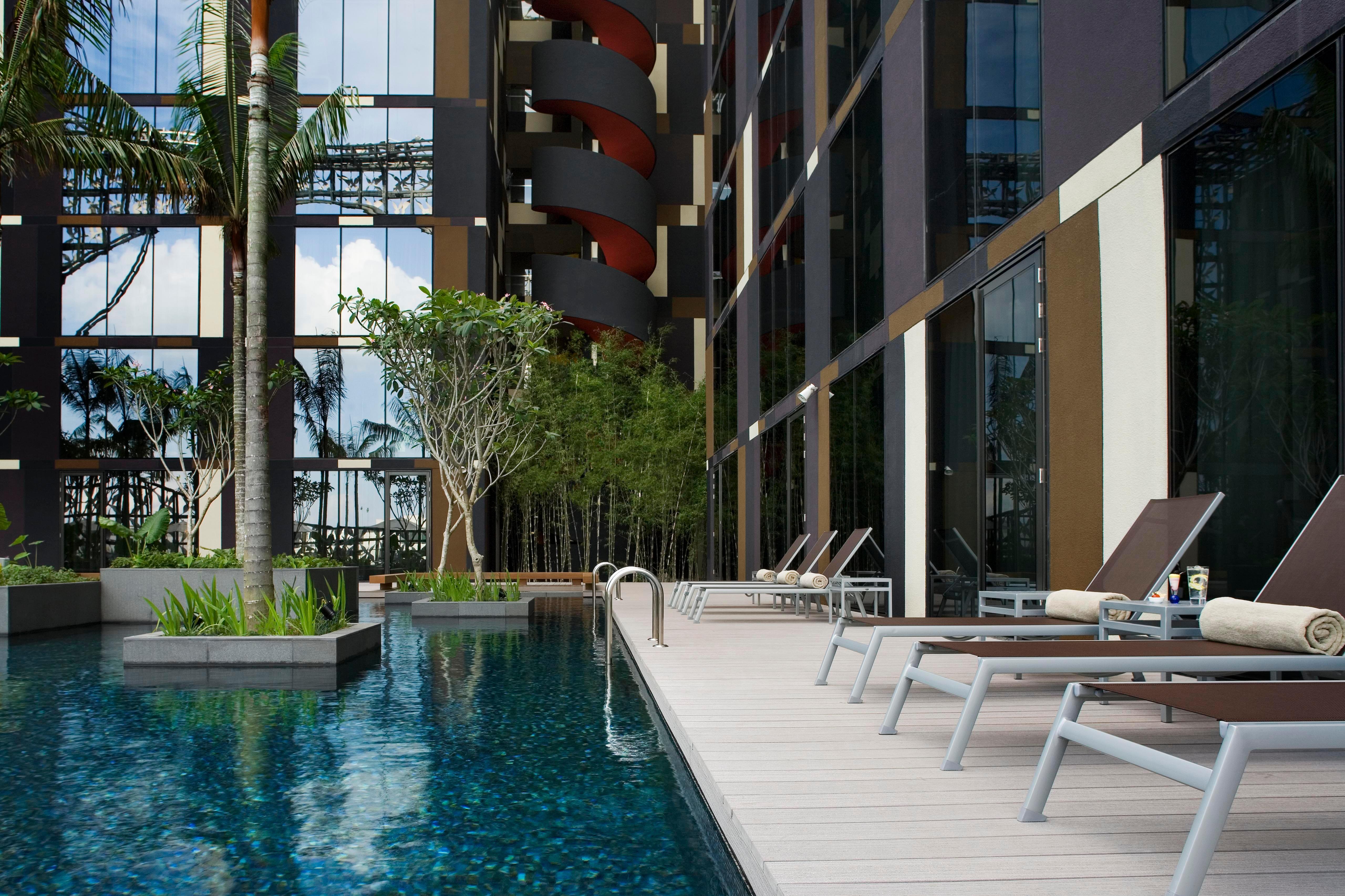 Crowne Plaza Changi Airport By Ihg Σιγκαπούρη Ανέσεις φωτογραφία The photo shows a modern swimming pool area adjacent to a building. The pool is surrounded by sleek wooden decking and features a clear blue water surface. There are several lounge chairs positioned around the pool for relaxation. Lush greenery, incl
