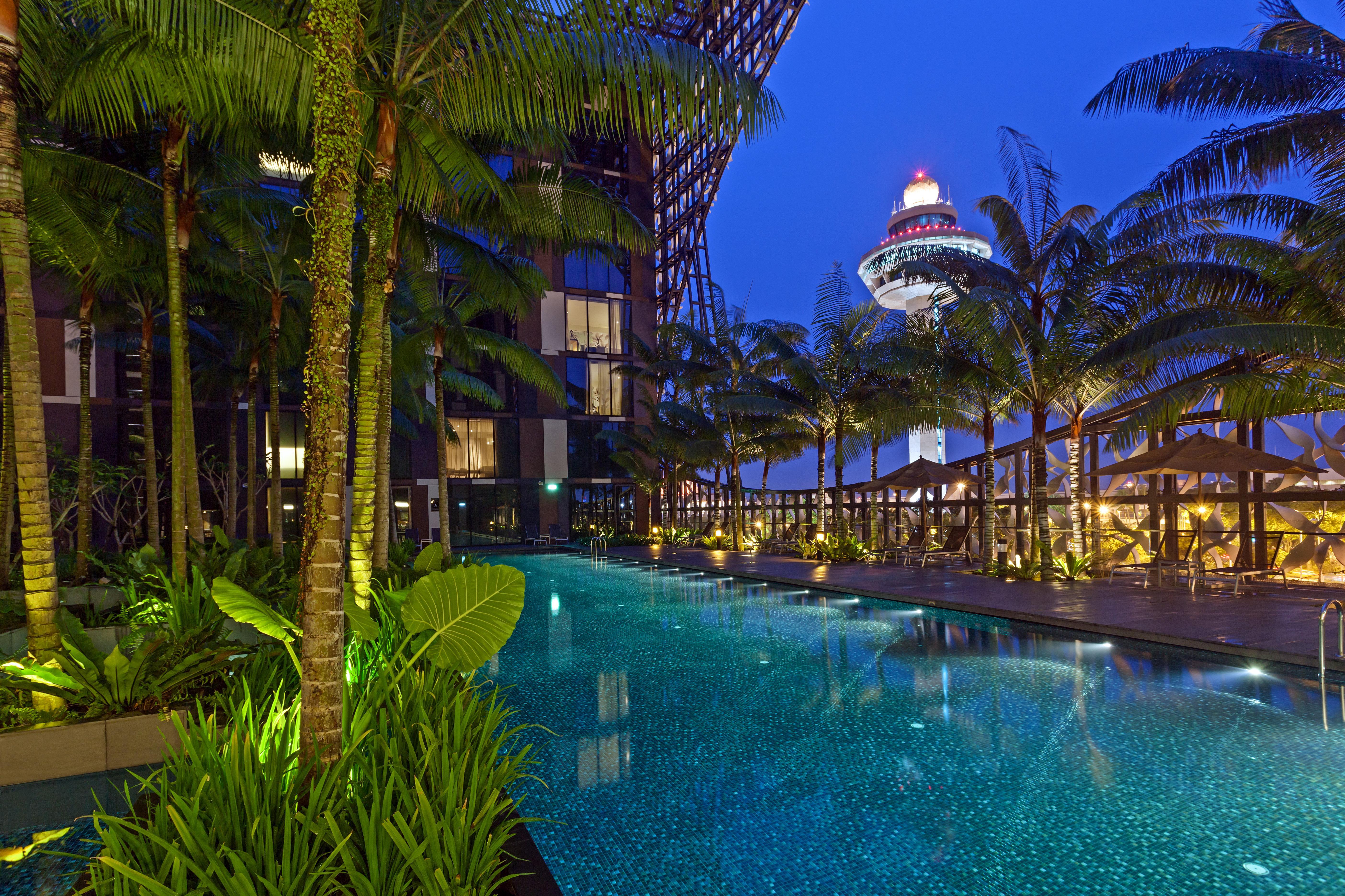 Crowne Plaza Changi Airport By Ihg Σιγκαπούρη Εξωτερικό φωτογραφία The photo depicts a luxurious outdoor swimming pool surrounded by lush palm trees and vibrant greenery. The scene is set during the evening, with soft lighting illuminating the water and plants. In the background, you can see a modern building struct