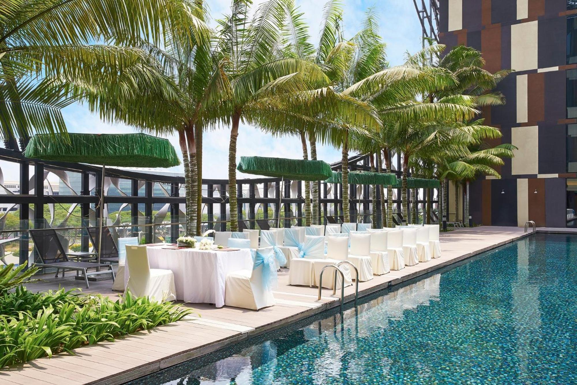 Crowne Plaza Changi Airport By Ihg Σιγκαπούρη Εξωτερικό φωτογραφία The photo shows a swimming pool area with a modern design. There are several palm trees lining the poolside, creating a tropical ambiance. Alongside the pool, there are white chairs and tables set up, possibly for an outdoor dining or event space. Th