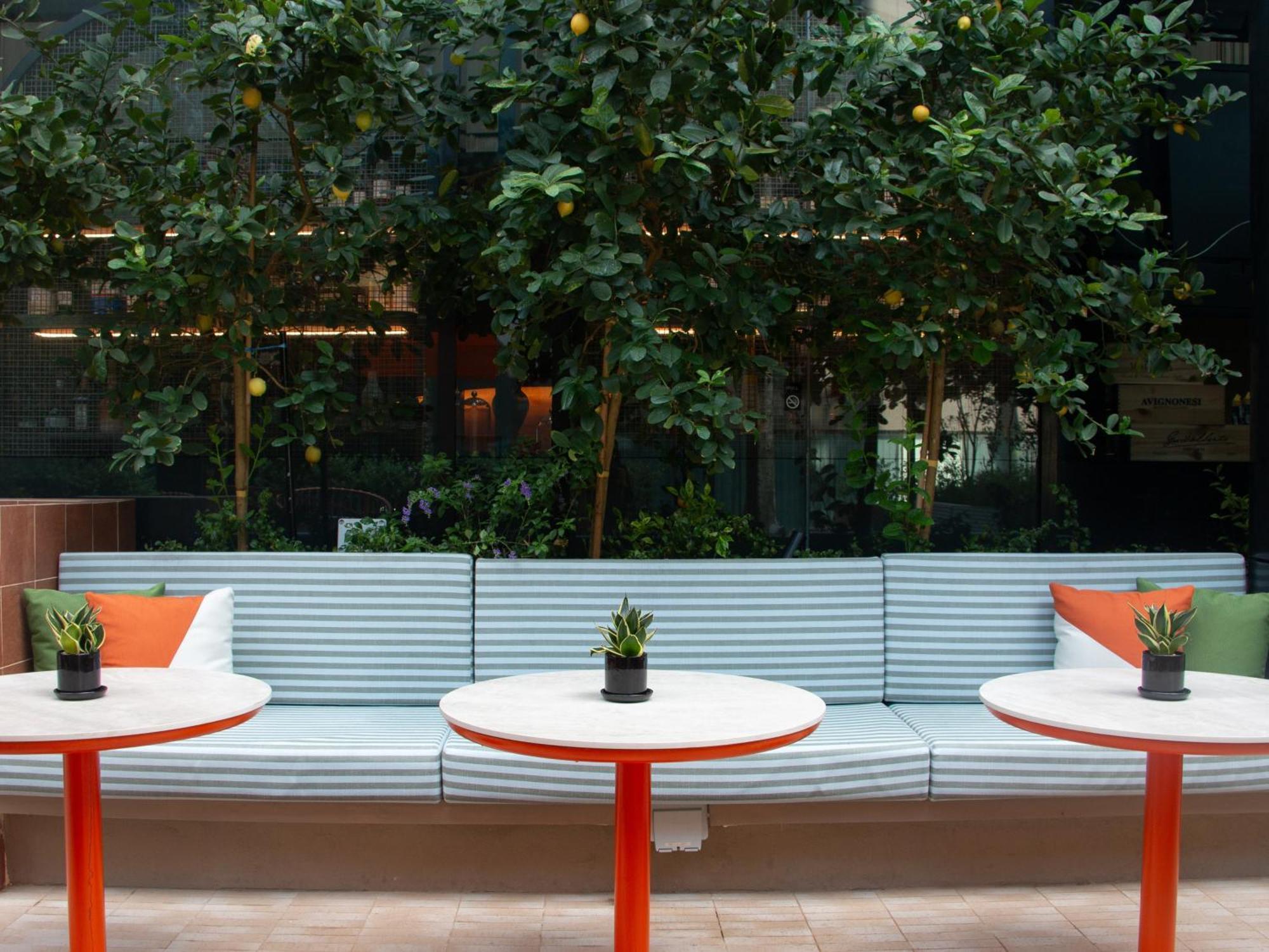 Crowne Plaza Changi Airport By Ihg Σιγκαπούρη Εξωτερικό φωτογραφία The photo shows a cozy seating area featuring a long, light blue bench with colorful cushions in orange and green. In front of the bench, there are three round tables with bright red bases and small plants in black pots on each table. The background 
