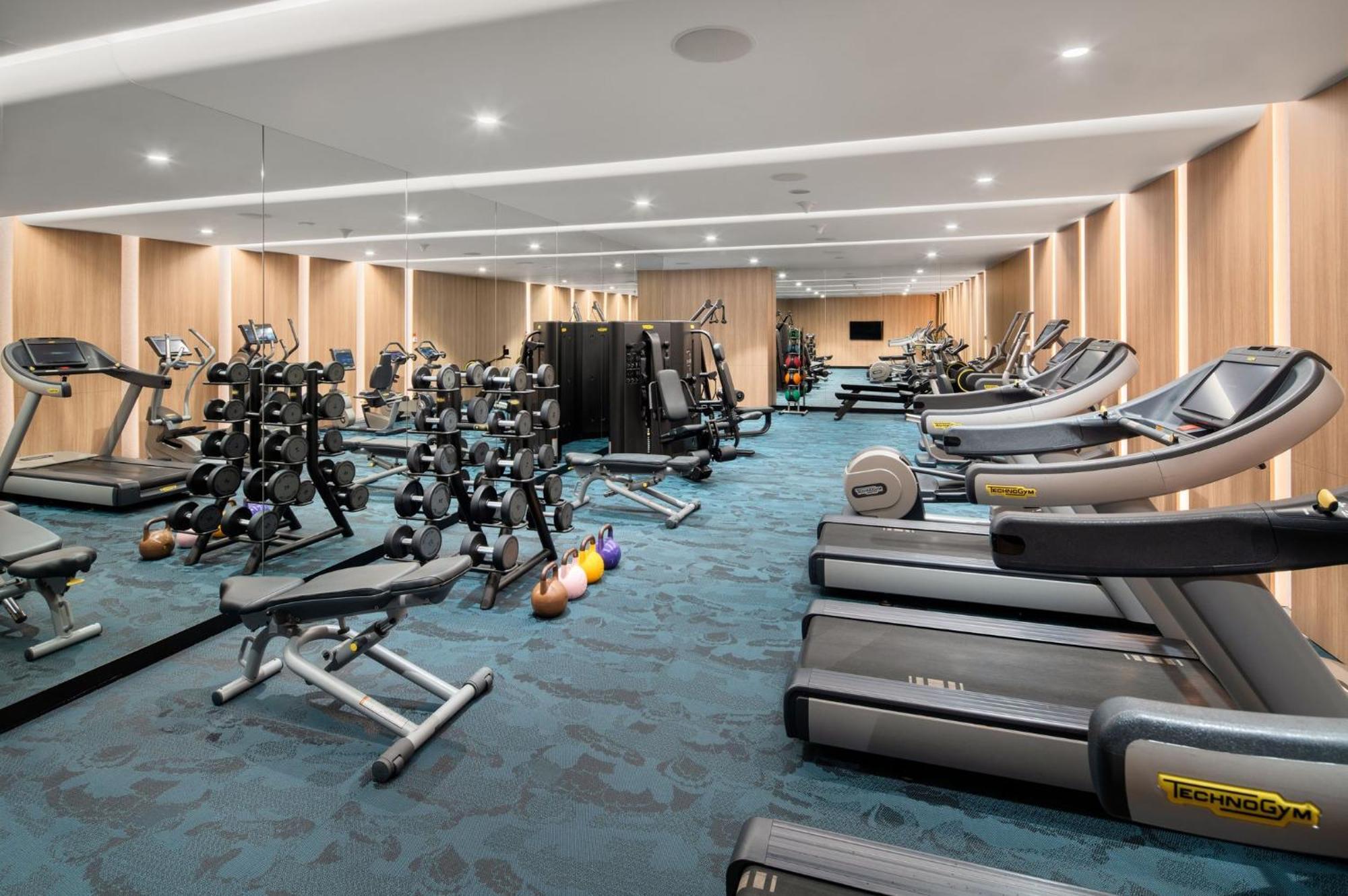 Crowne Plaza Changi Airport By Ihg Σιγκαπούρη Εξωτερικό φωτογραφία The photo shows a modern gym interior. It features various fitness equipment, including multiple treadmills, weightlifting machines, and a set of free weights on racks. The flooring has a blue carpet, and the walls are made of wood paneling, giving t
