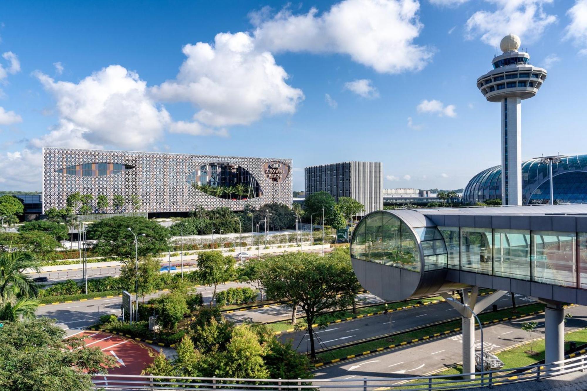 Crowne Plaza Changi Airport By Ihg Σιγκαπούρη Εξωτερικό φωτογραφία The photo shows a modern urban landscape with a clear blue sky and fluffy white clouds. In the foreground, there is a landscaped area with greenery and a road. Prominently featured is an interesting architectural structure, likely a building with a u